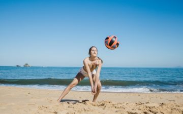 Esportes e atividades para praticar na praia: vôlei. Na imagem, uma jovem está na praia posicionada para uma manchete na bola de vôlei