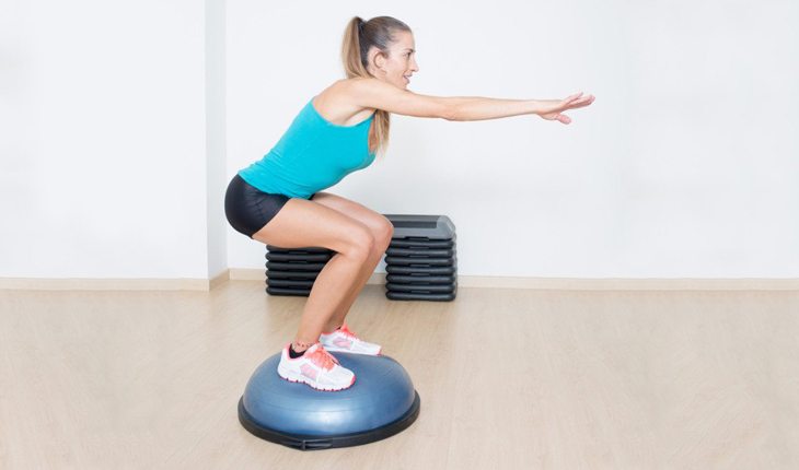 Imagem de uma mulher em uma academia realizando um agachamento com um bosu. Bosu