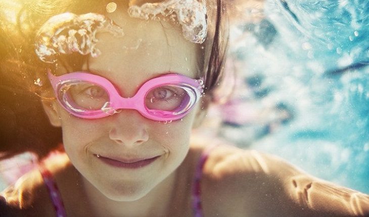 a foto mostra uma menina sorrindo ambaixo da água com um óculos de natação rosa