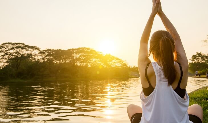 Mulher sentada com os braços para o alto, encarando o pôr do sol. Estresse pode atrapalhar o processo de emagrecimento