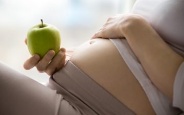 Alimentação na gravidez: na foto, a barriga de uma mulher grávida parcialmente à mostra, enquanto ela segura uma maçã com a mão apoiada no ventre