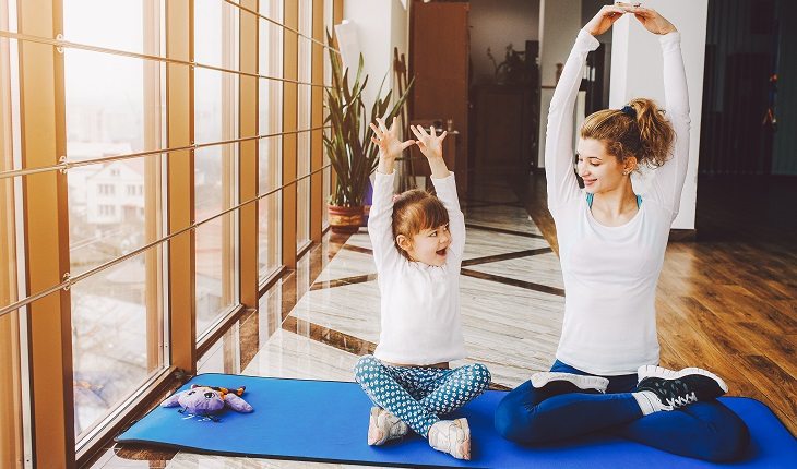 Mãe e filha sentadas em um colchonete com os braços para cima fazendo exercício