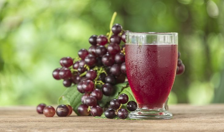 A foto mostra um cacho de uma ao lado de um copo com suco de uva, um dos alimentos antienvelhecimento