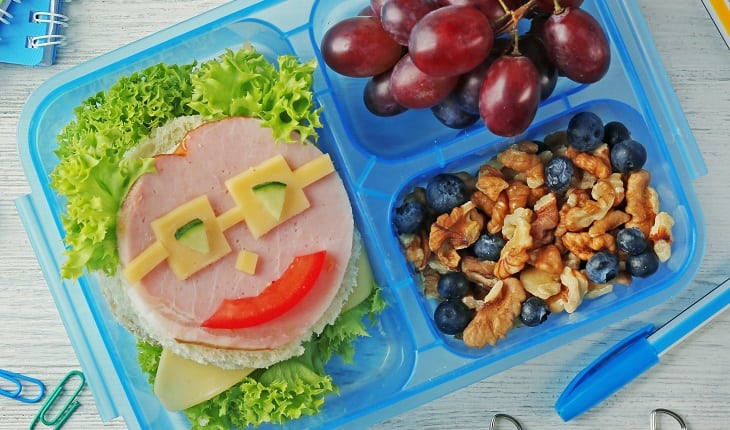 Alimentos nutritivos para as crianças: dicas para as férias de julho. Na foto, uma lancheira com alimentos coloridos e um lanche com carinha.