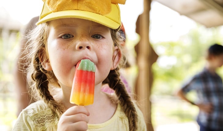 Alimentos nutritivos para as crianças: dicas para as férias de julho. Na foto, uma menininha chupando um picolé colorido