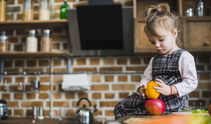 Alimentação vegana na gravidez: na foto, uma criança sentada em uma bancada de cozinha mexendo e olhando para duas frutas.