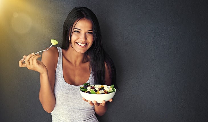 A foto mostra uma mulher sorrindo comendo uma refeição equilibrada com muita salada