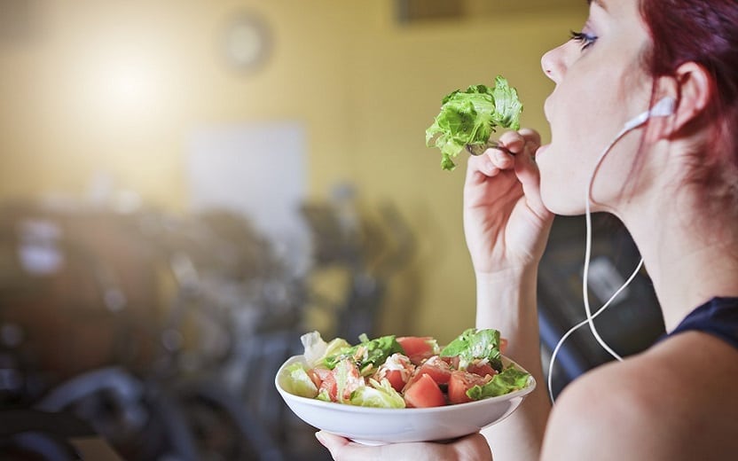 A foto mostra uma mulher com fone de ouvido comendo salada em uma academia. A foto ilustra o tema do post sobre indicações de alimentos para corredores