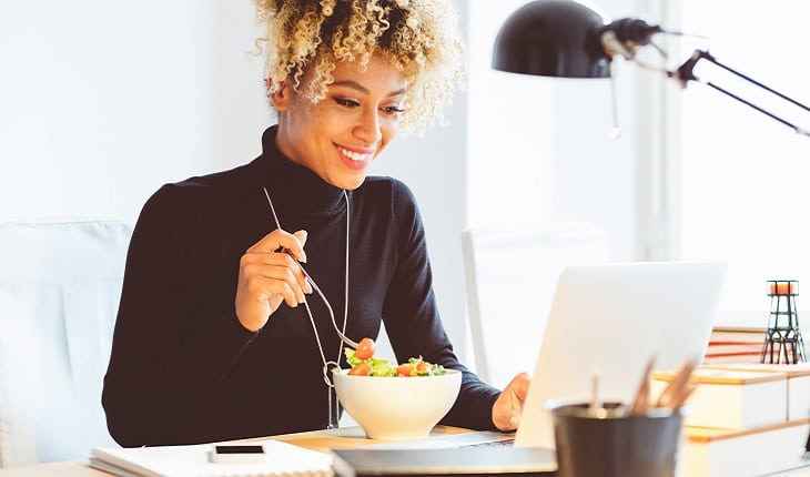 A foto mostra uma mulher tomando café da manhã enquanto trabalha