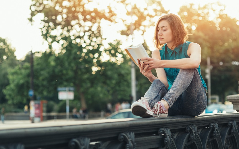 Garota sentada ao ar livre lê um livro e coloca em prática os ensinamentos sobre a importância da atividade física para os estudos