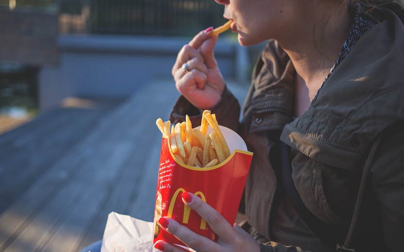 Uma mulher se alimenta de batatas fritas. Ela as segura, aparentemente sentada. Ao fundo, um piso de madeira com alguns feixes de sol iluminando o chão. O nutriente é um comfort food