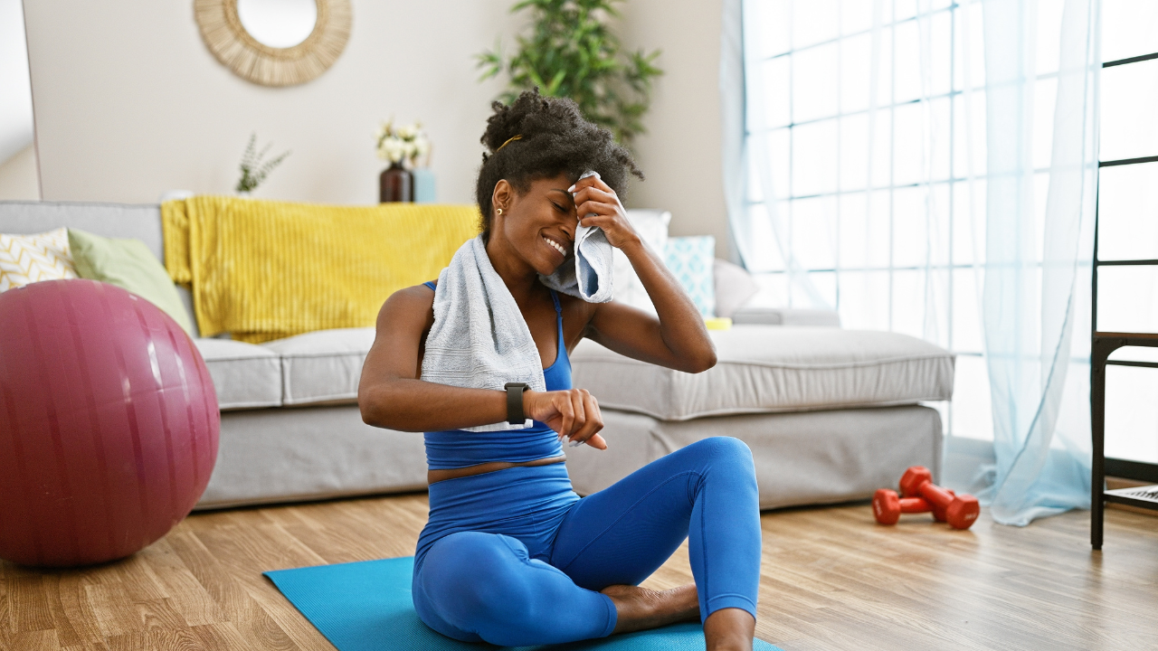 Mulher jovem descansando após treino em casa, sentada no tapete de exercícios e enxugando o suor, com halteres e bola de pilates ao fundo.