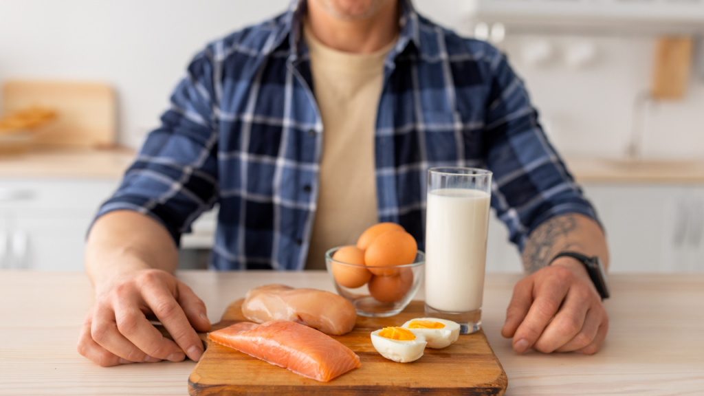 A imagem mostra um homem segurando uma bandeja com alimentos: copo de leite, salmão, ovos e peito de frango.