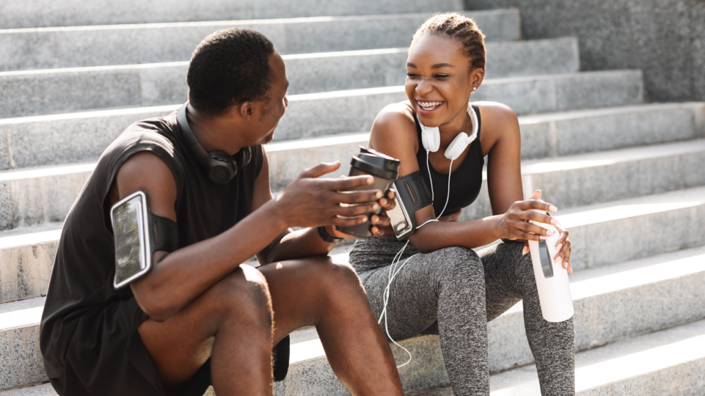 A imagem mostra um homem negro e uma mulher negra sentados em uma escada ao ar livre conversando e sorrindo. Eles vestem roupas de exercícios físico.