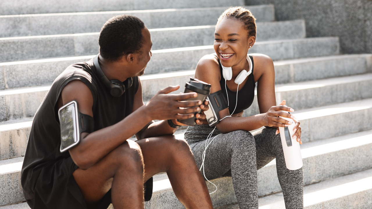 A imagem mostra um homem negro e uma mulher negra sentados em uma escada ao ar livre conversando e sorrindo. Eles vestem roupas de exercícios físico.