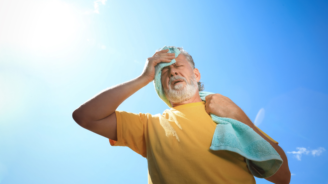 Imagem de um homem idoso secando o suor com uma toalha azul enquanto caminha em um dia de sol