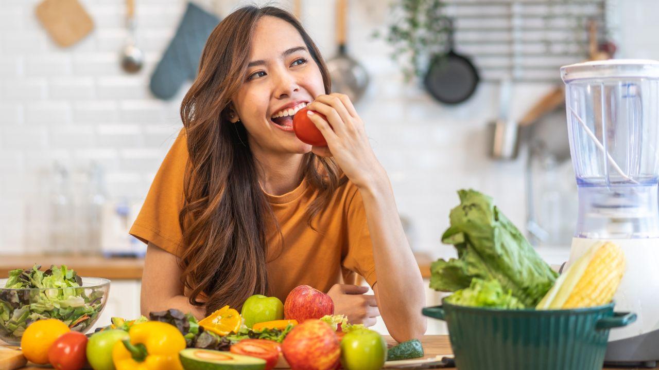 Mulher branca, cabelo liso longo, mordendo uma maça sorrindo. Na frente dela, tem uma mesa repleta de frutas e vegetais.