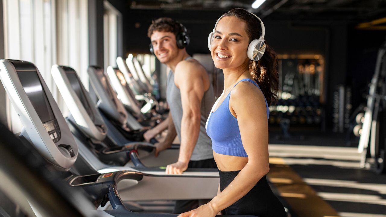 Mulher na esteira da academia sorrindo para a câmera e usando fones de ouvido.