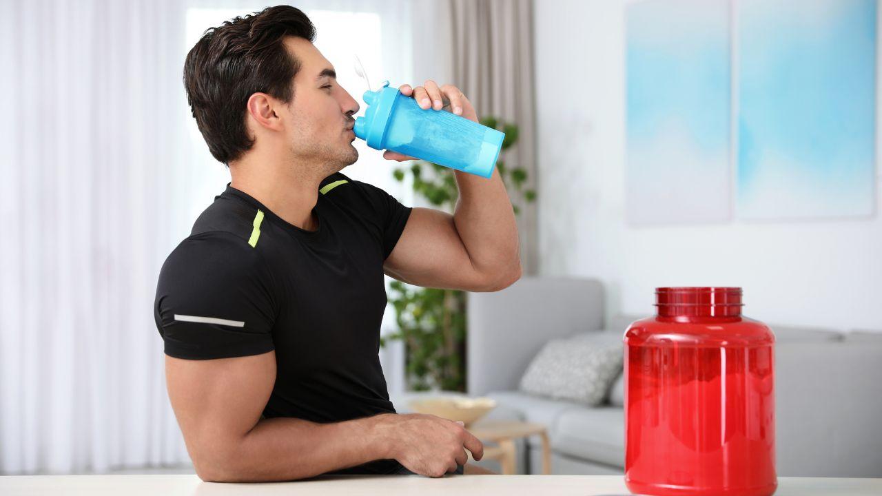 Homem branco, camiseta preta, com braço malhado tomando whey protein.