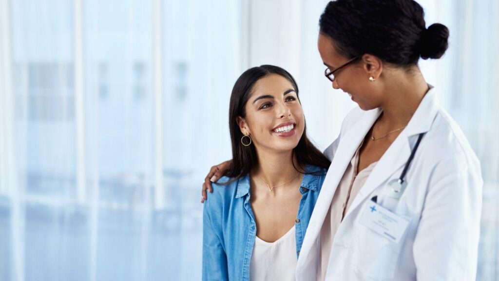 Mulher branca sorrindo e abraçando médica negra.