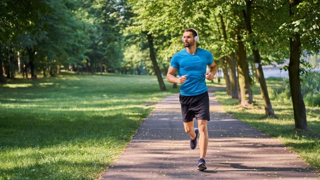 Homem branco, cabelo castanho, barba, usando camiseta azul e short preto correndo num parque ao ar livre.