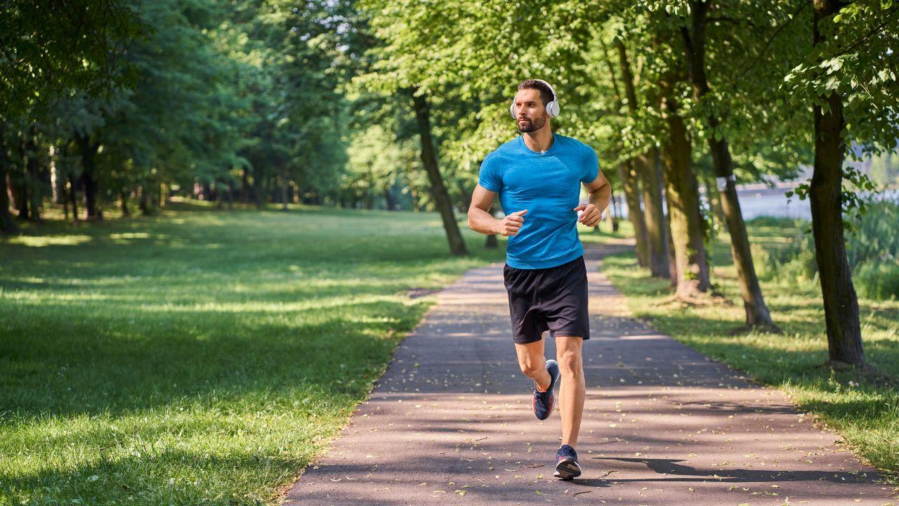 Homem branco, cabelo castanho, barba, usando camiseta azul e short preto correndo num parque ao ar livre.