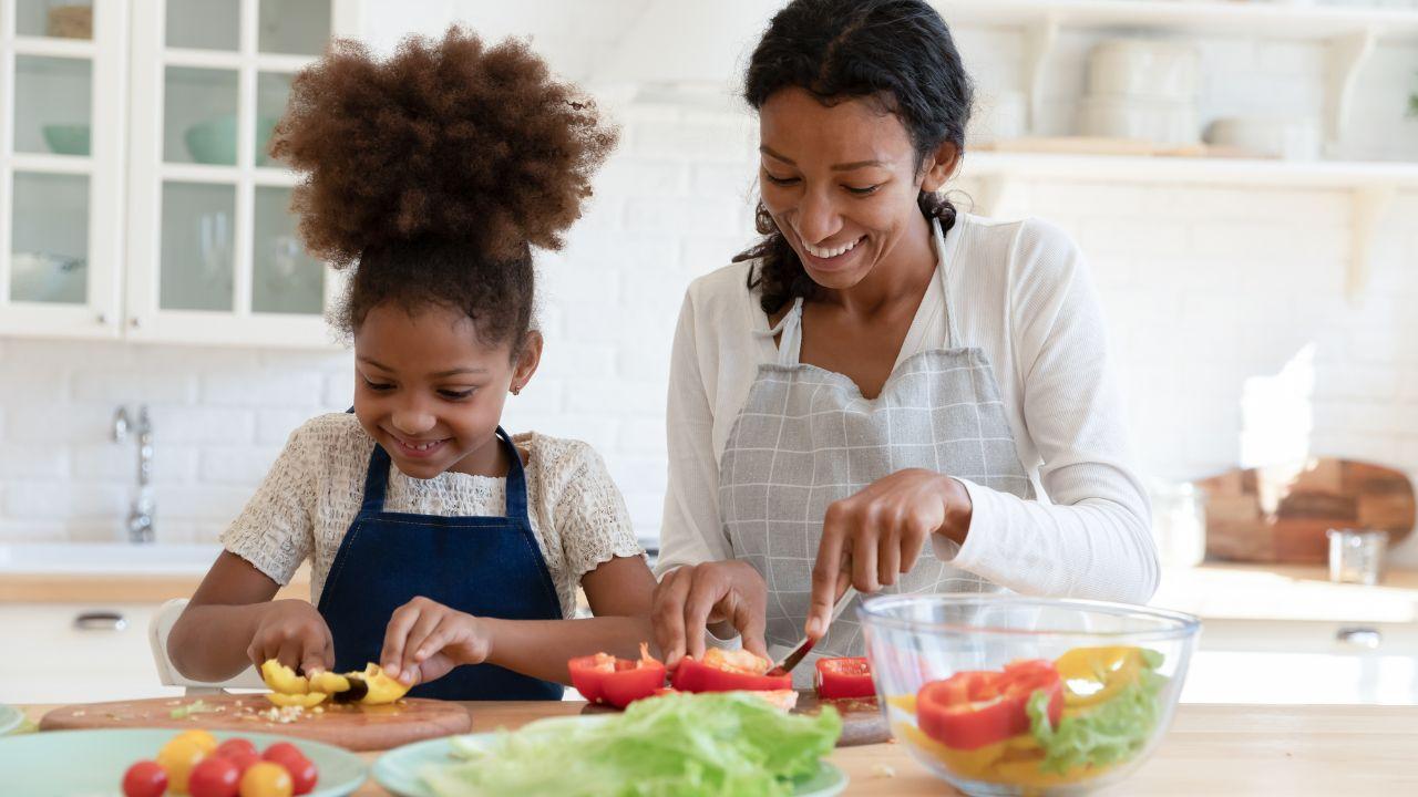 Mulher e criança negra cortando legumes em uma bancada na cozinha.