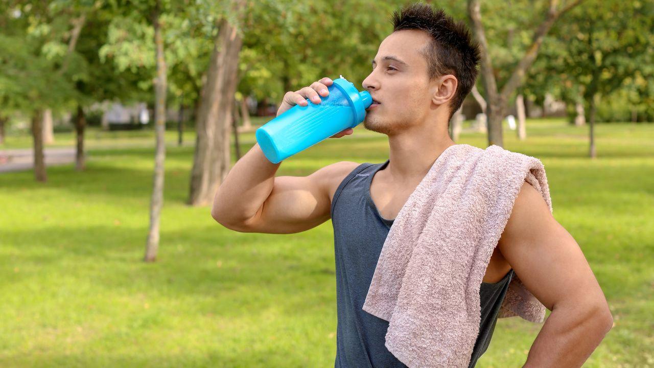 Homem com regata e toalha no ombro, bebendo alguma coisa na garrafa de whey.