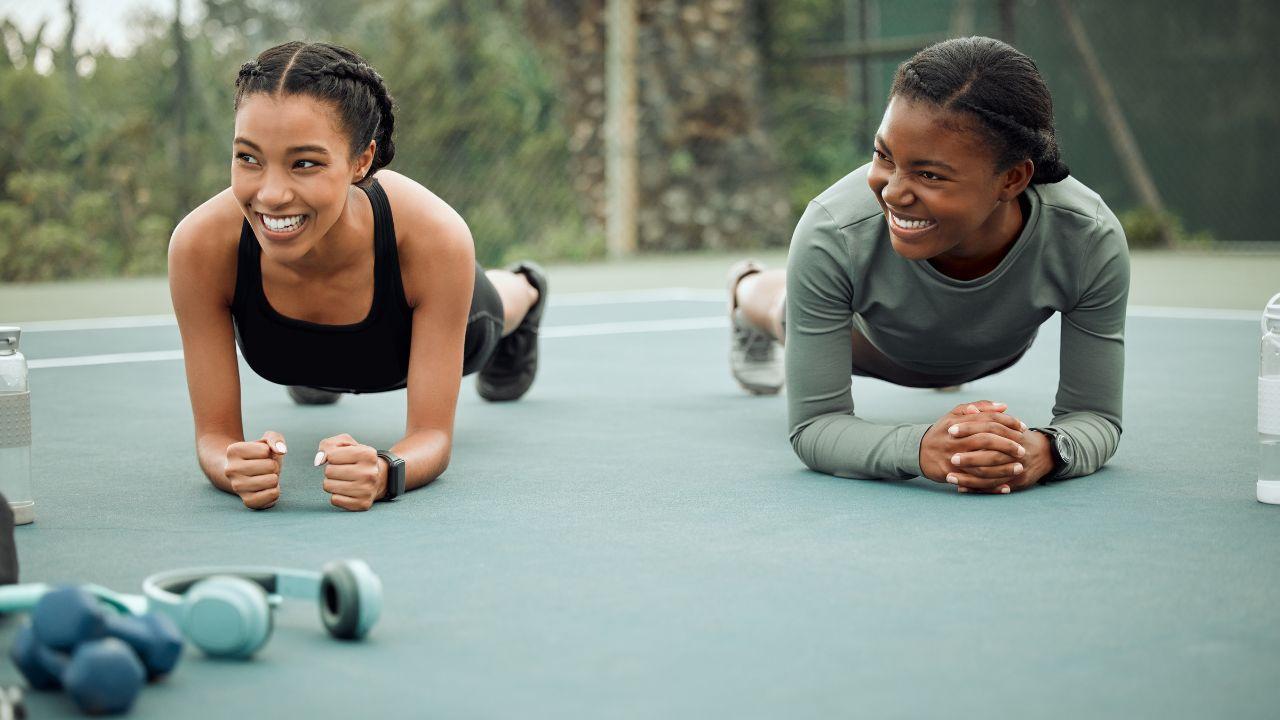 Duas mulheres negras fazendo prancha em um colchonete ao ar livre. Elas sorriem.