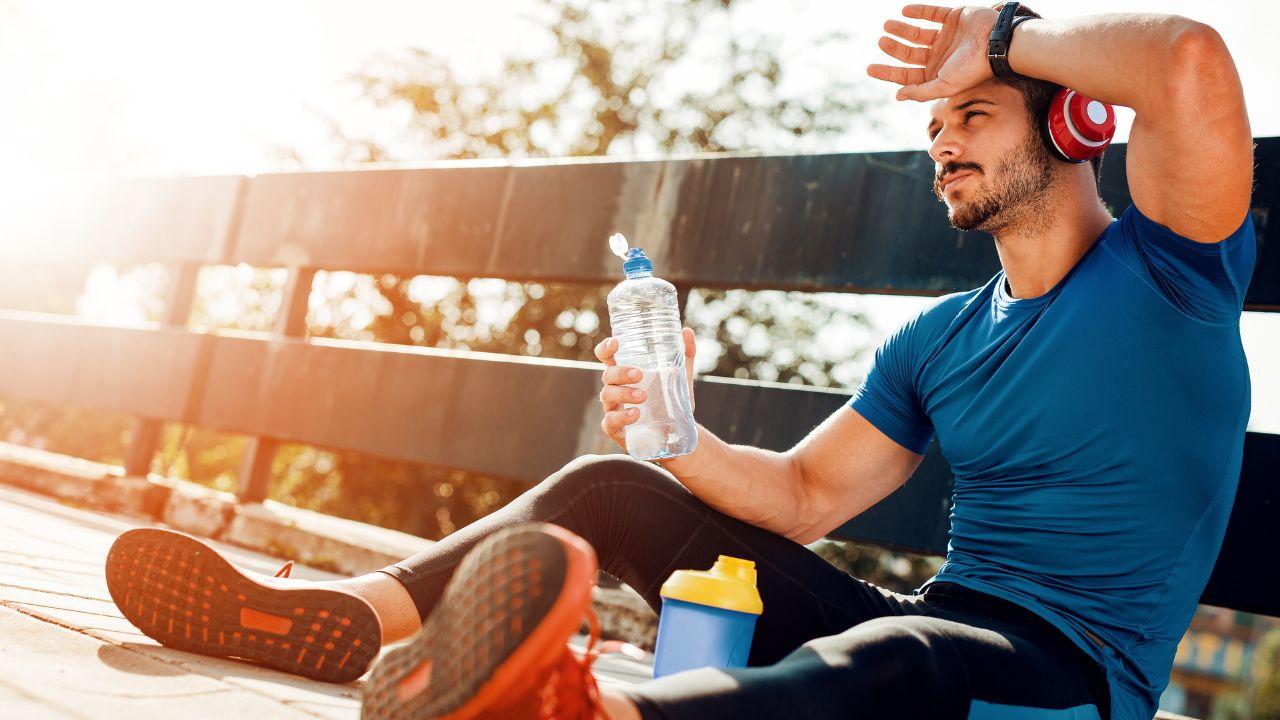 Imagem de um homem com roupa de corrida sentado no chão, com o braço na testa se protegendo dos raios de sol. Ele segura uma garrafinha em uma das mãos