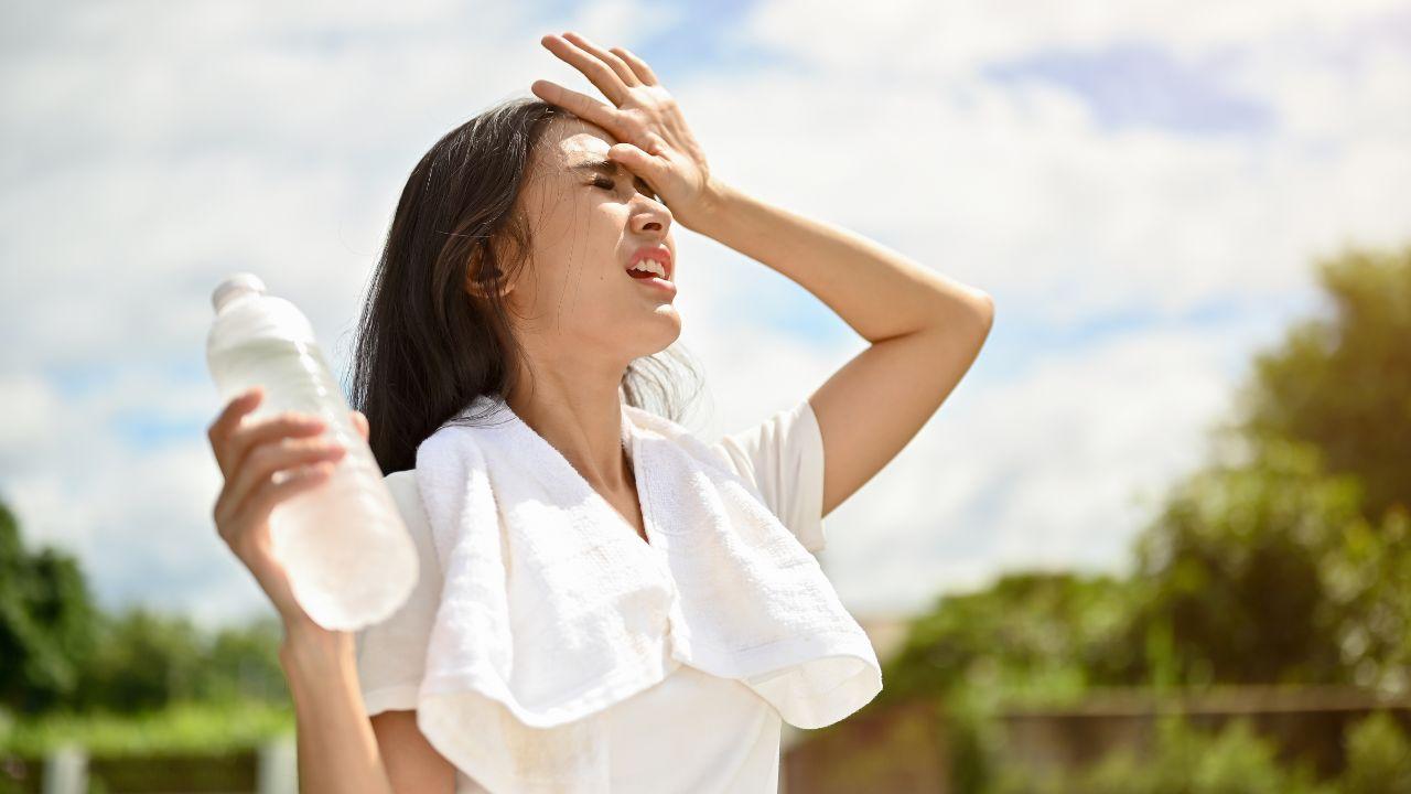 Imagem de uma mulher suando, com a mão na testa, uma garrafinha na outra mão e uma toalha branca sobre os ombros, em um dia quente enquanto faz exercícios físicos