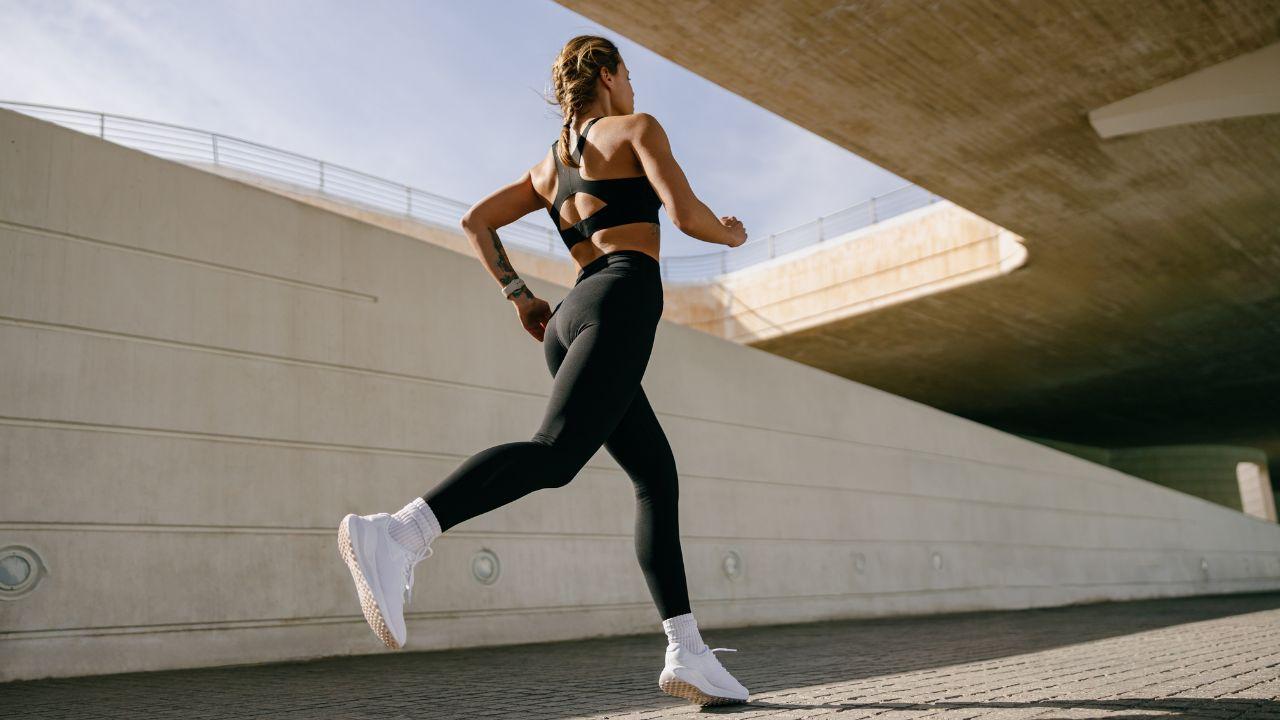 Imagem de uma mulher, de costas, correndo. Ela veste um top com recorte na costas e legging, ambos pretos; além de um tênis branco