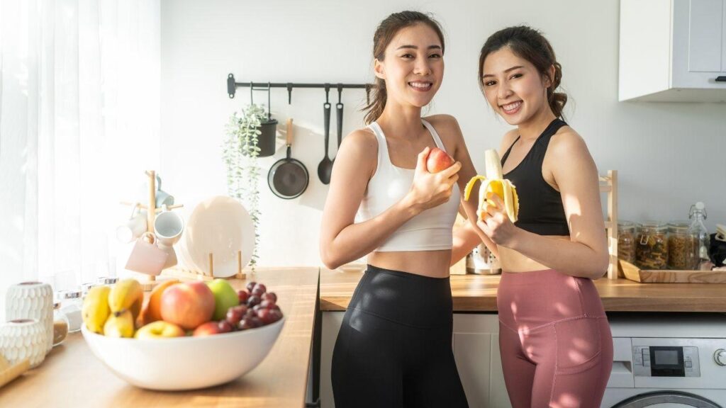 Imagem de duas mulheres, vestindo roupa de academia, na cozinha; a da direita segura uma banana e a da esquerda uma maçã. Há uma fruteira com frutas diversas na bancada ao lado