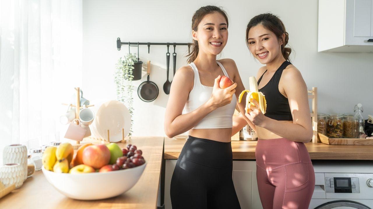 Imagem de duas mulheres, vestindo roupa de academia, na cozinha; a da direita segura uma banana e a da esquerda uma maçã. Há uma fruteira com frutas diversas na bancada ao lado
