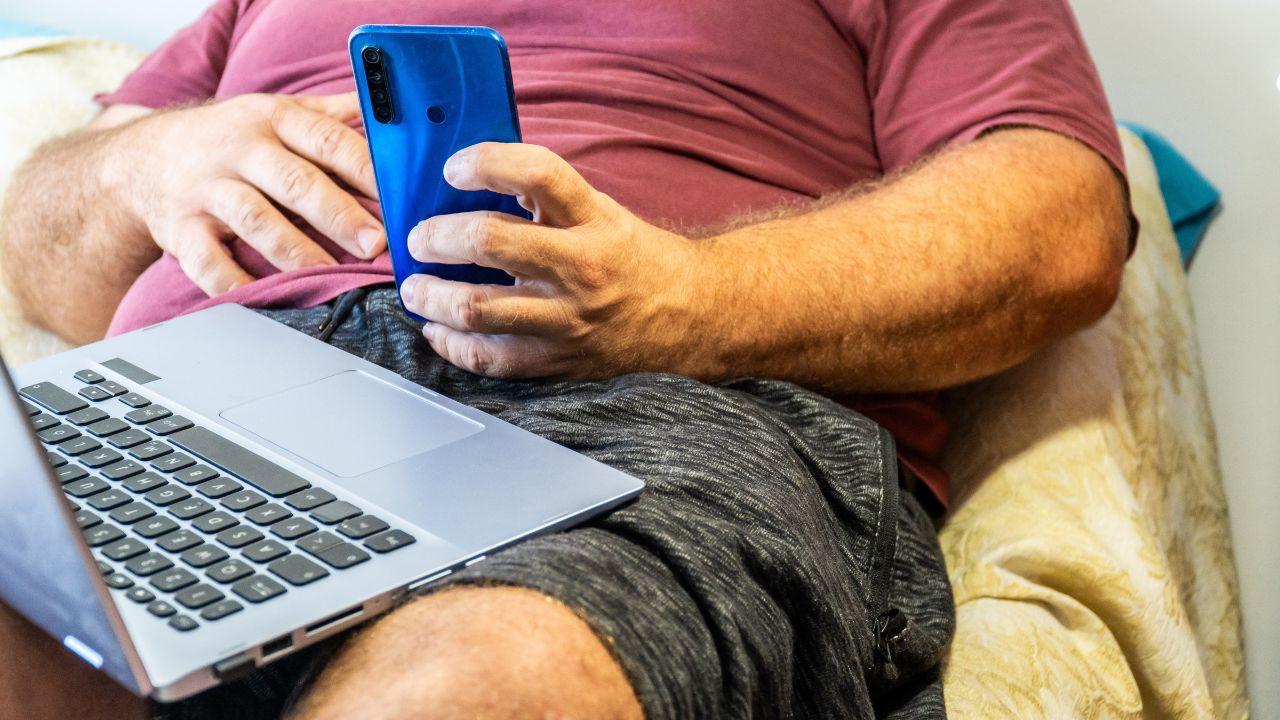 Imagem de uma pessoa obesa no sofá, mexendo no celular e no computador