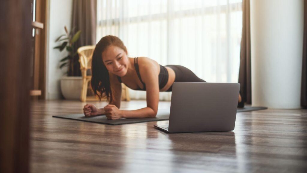 Mulher na posição de prancha no colchonete na frente de um computador.
