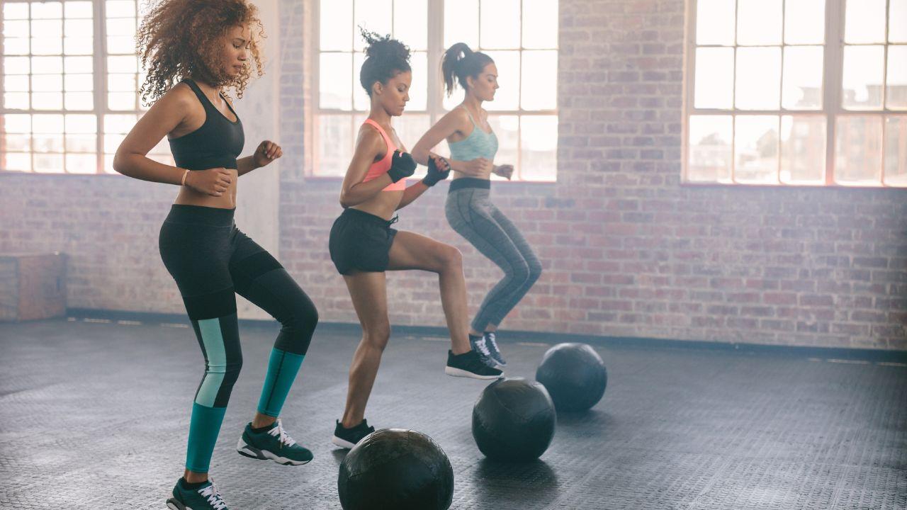 Mulheres fazendo exercício com bola no chão da academia.