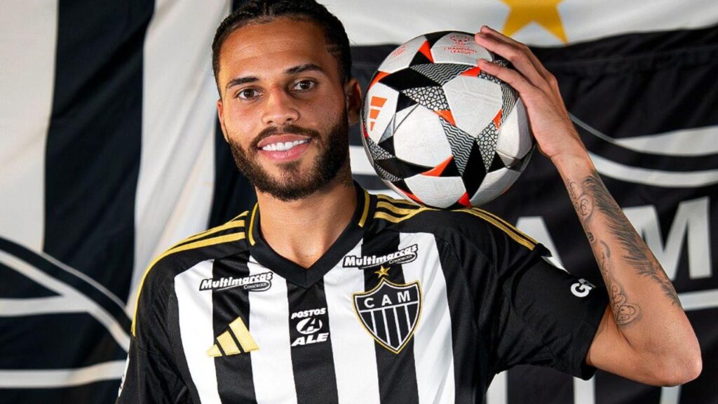 Jogador Alexsander. Homem negro, cabelo curto preto, uniforme do Atlético mineiro e ele segurando uma bola no ombro.