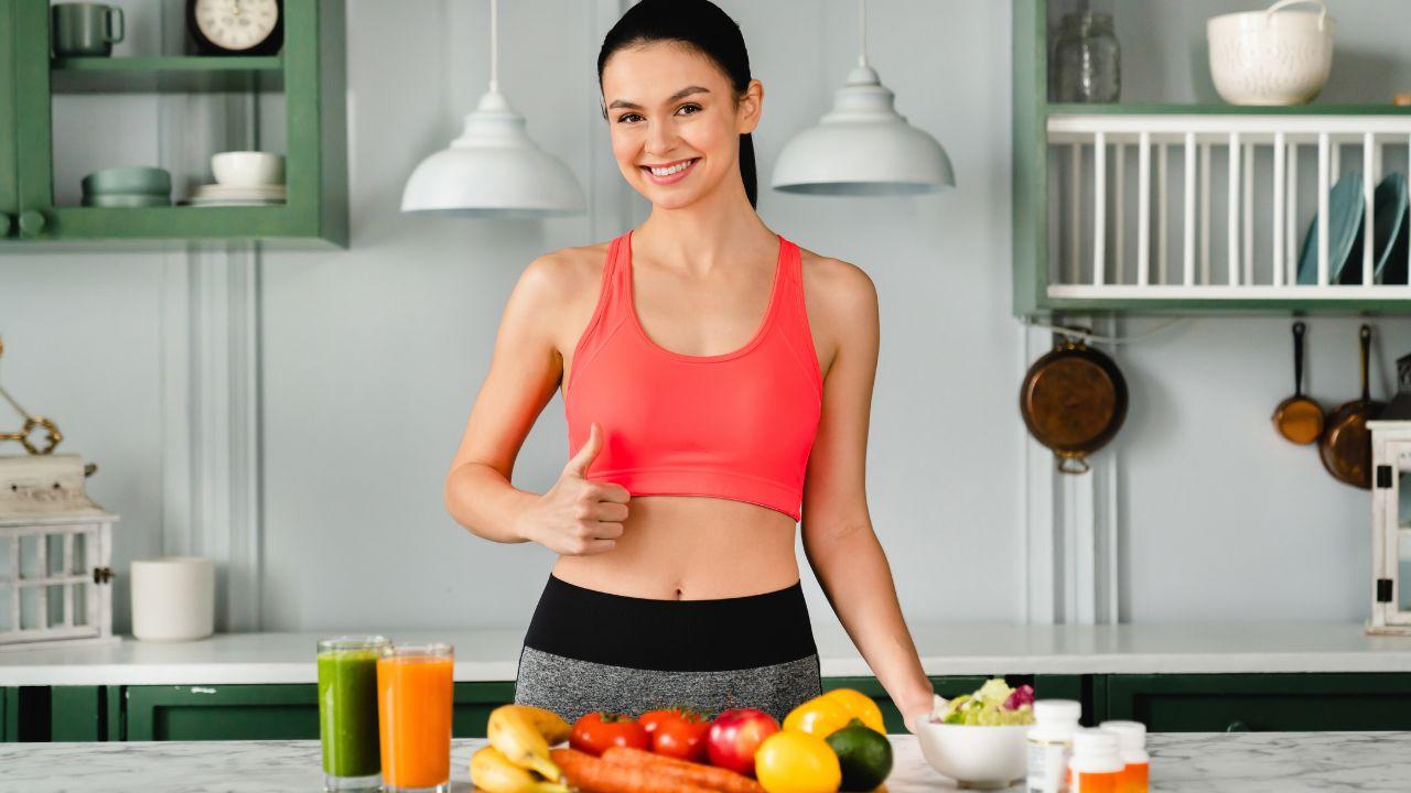 Mulher com roupa fitness fazendo um joinha na frente de uma mesa com alimentos saudáveis como frutas e verduras.