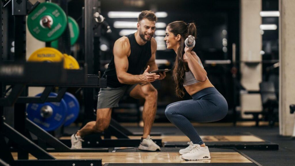Imagem de uma mulher fazendo agachamento com barra nas costas e personal do lado