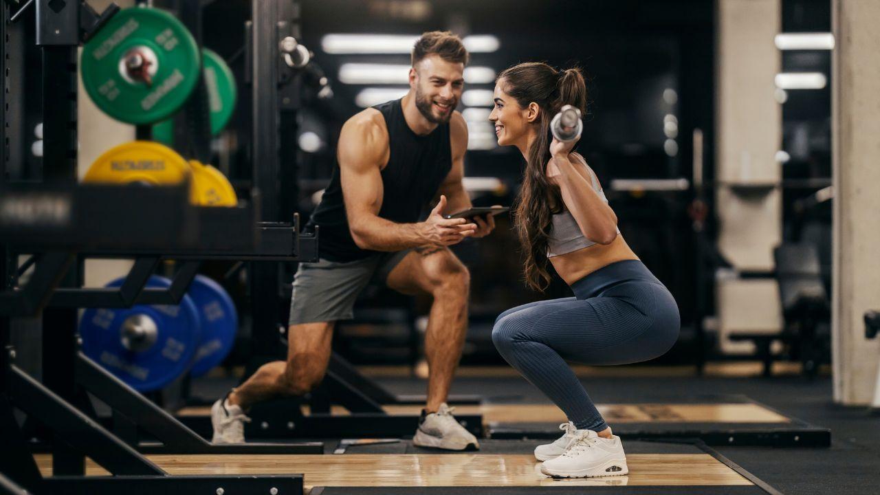 Imagem de uma mulher fazendo agachamento com barra nas costas e personal do lado