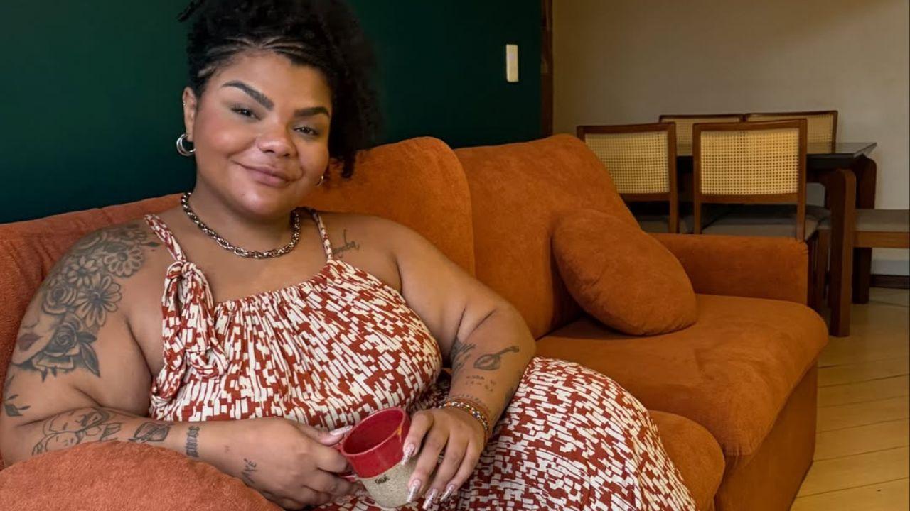 Flora Cruz na foto. Mulher negra, cabelo cacheado preso, sentada num sofá, usando vestido longo florido.