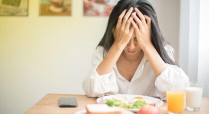 Mulher sentada na mesa de café da manhã com as mãos na cabeça com feição preocupada.