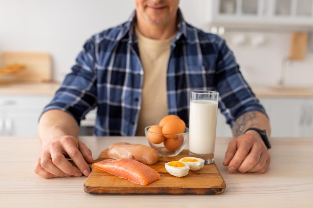 Foto mostra homem sentado na mesa com um prato com ovos, leite e peito de frango na frente.