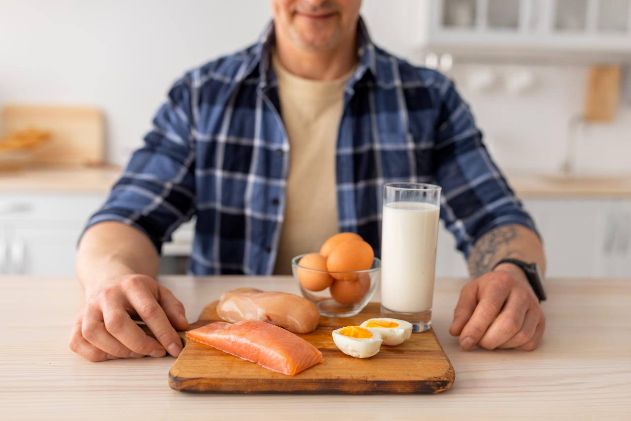 Foto mostra homem sentado na mesa com um prato com ovos, leite e peito de frango na frente.