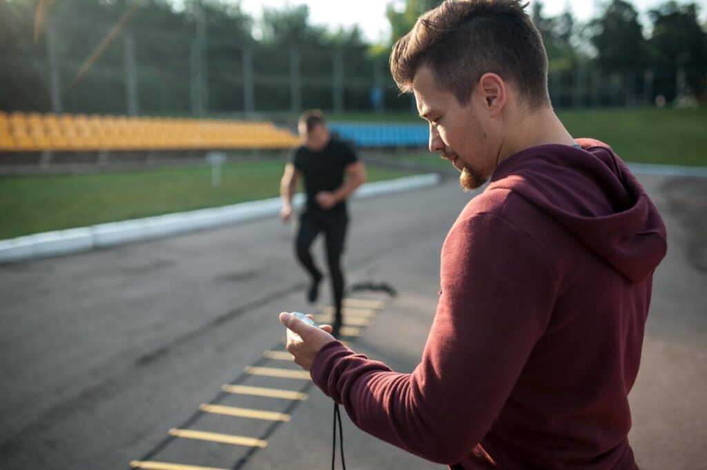 Foto mostra homem olhando pro relógio enquanto outro homem faz exercício com cordas.