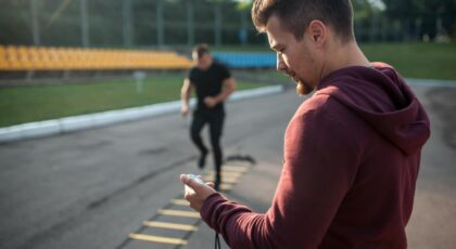 Foto mostra homem olhando pro relógio enquanto outro homem faz exercício com cordas.