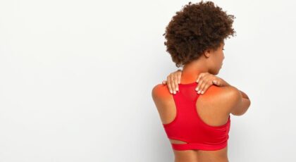 Imagem de uma mulher com roupa de treino e as mãos na região dos ombros, que está em vermelho, sentindo dor