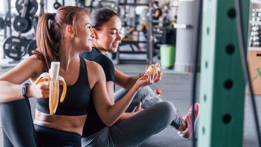 Imagem duas mulheres na academia, uma comendo uma banana enquanto entrega um lanche para a amiga