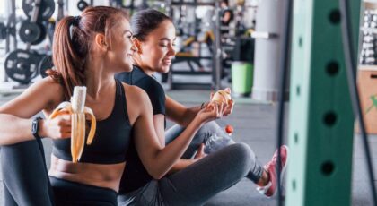 Imagem duas mulheres na academia, uma comendo uma banana enquanto entrega um lanche para a amiga
